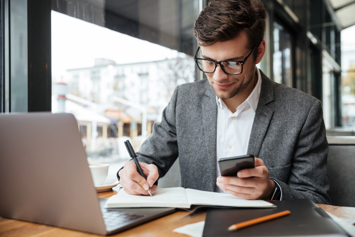 smiling businessman eyeglasses sitting by table cafe with laptop computer while using smartphone writing something blauw licht filter bril man
